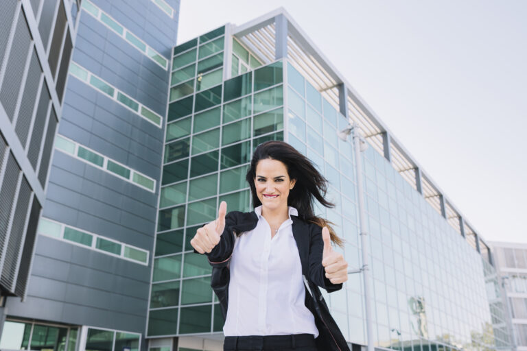 Mulher de negócios sorridente com gesto de positivo em frente a um prédio moderno.